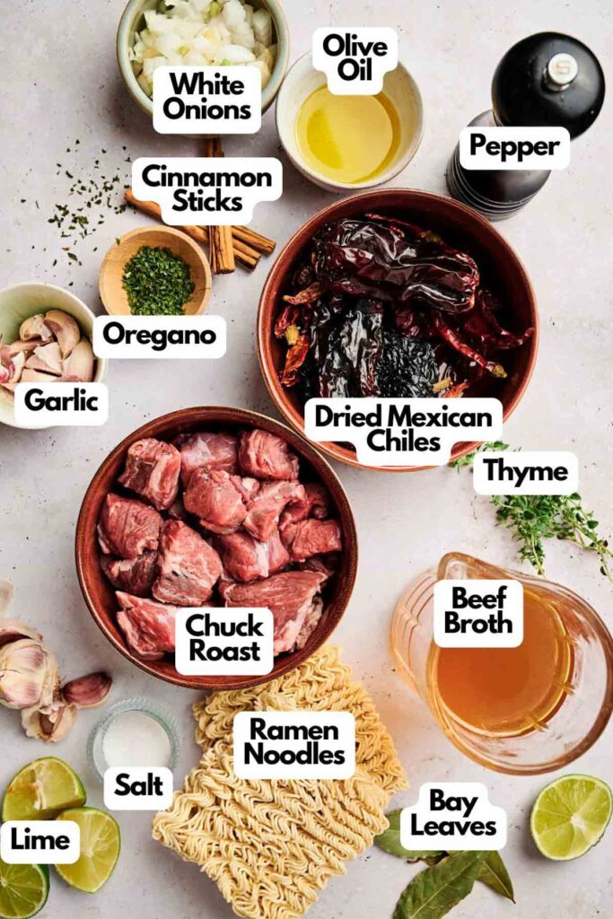 Various ingredients labeled for a birria ramen recipe, including chuck roast, ramen noodles, dried Mexican chiles, beef broth, and spices like oregano and thyme, are artfully arranged on a light surface.