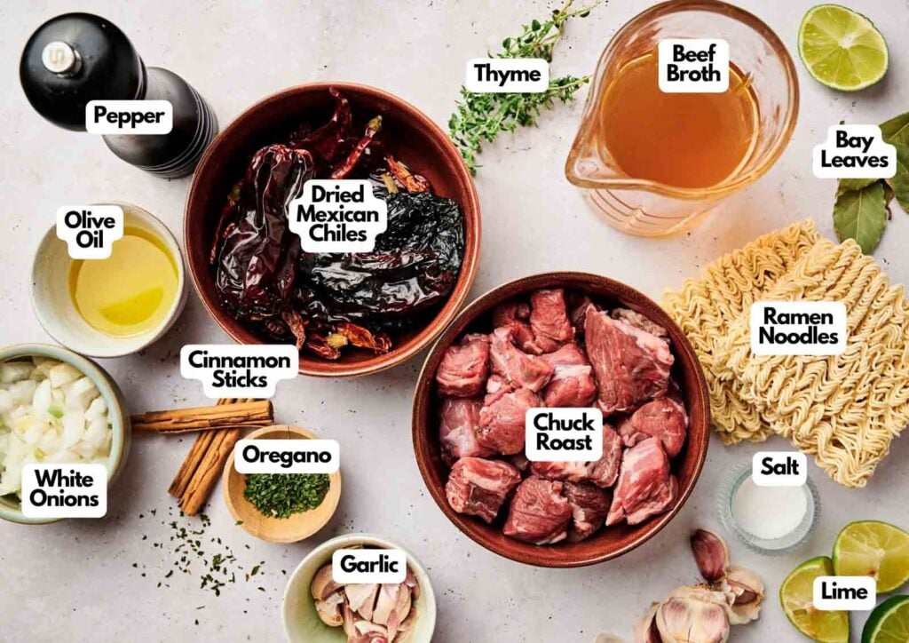 Various labeled ingredients for birria ramen are arranged on a countertop, including chuck roast, dried Mexican chiles, ramen noodles, spices, broth, onions, garlic, and lime.