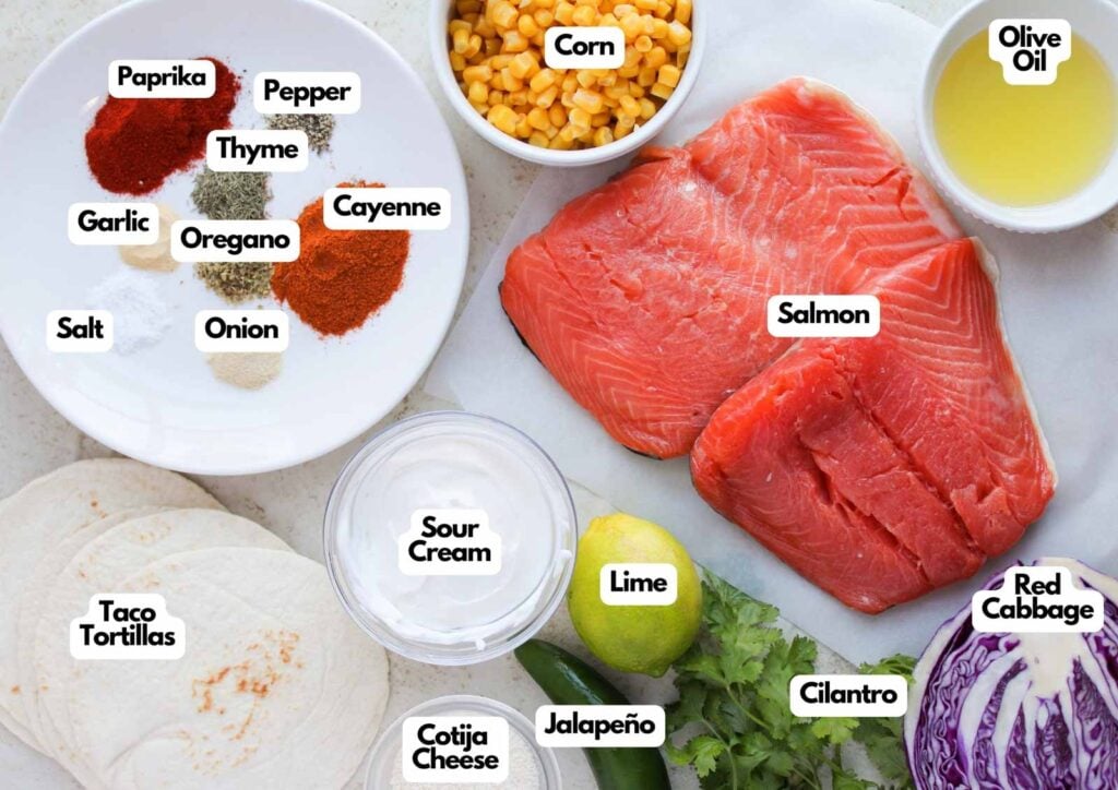 Ingredients for Blackened Salmon Tacos are arranged on a table, including raw salmon, spices, corn, olive oil, red cabbage, lime, cilantro, sour cream, jalapeño, cheese, and tortillas.
