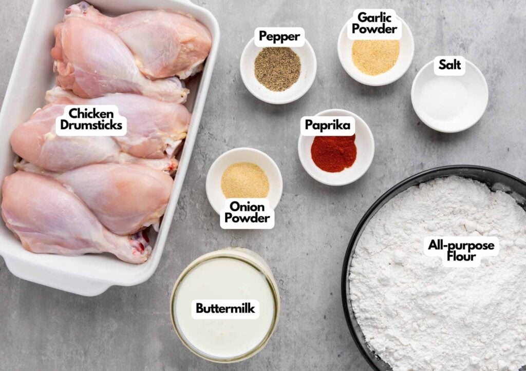 Raw chicken drumsticks in a dish, with bowls of all-purpose flour, buttermilk, salt, pepper, paprika, onion powder, and garlic powder on a countertop—everything you need for classic buttermilk fried chicken.