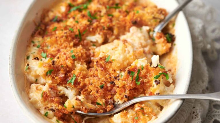 A dish of baked creamy cauliflower topped with golden breadcrumbs, garnished with chopped herbs, and served with two metal spoons.