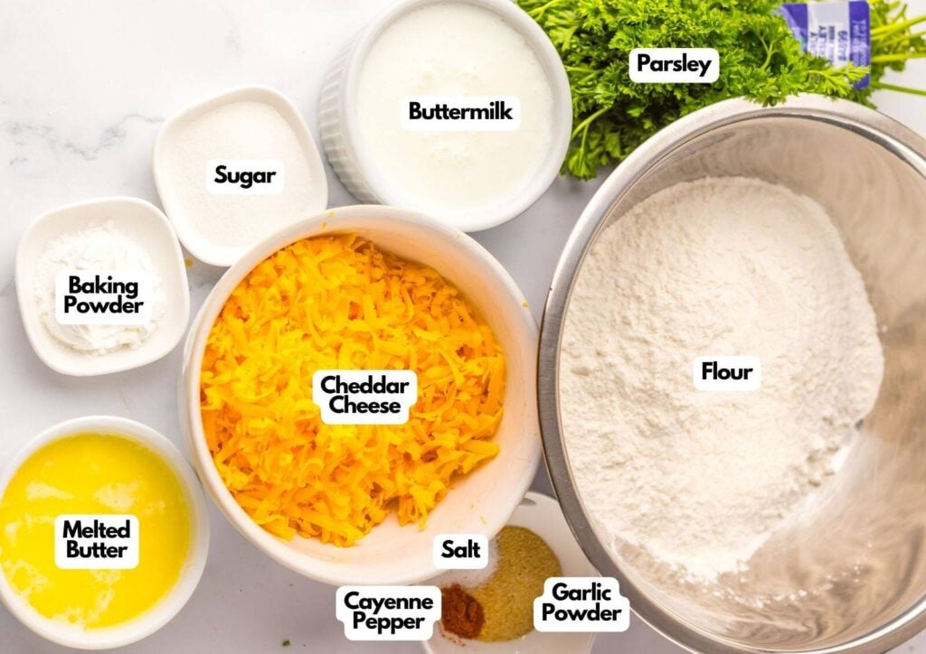 Various baking ingredients in labeled bowls, including flour, cheddar cheese, buttermilk, melted butter, parsley, sugar, baking powder, salt, cayenne pepper, and garlic powder—everything you need to make classic cheddar bay biscuits.