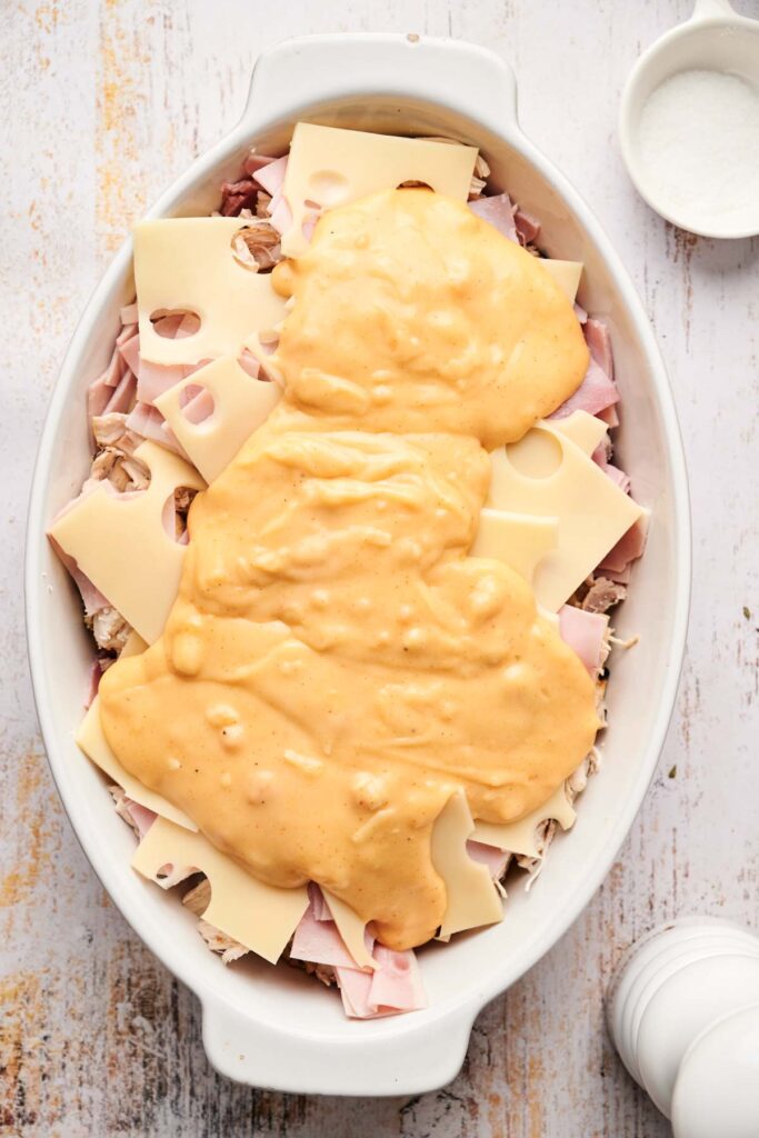 This Chicken Cordon Bleu Casserole recipe features layers of sliced cheese, ham, mushrooms, and a rich cheese sauce on top, elegantly arranged on a light wooden surface.