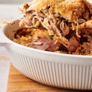 A serving of Chicken Cordon Bleu casserole with a breadcrumb topping is being lifted from a white dish on a wooden board.