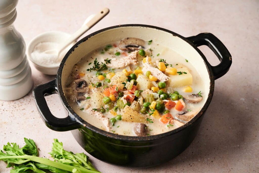 A pot of creamy chicken pot pie soup sits on a light surface, garnished with herbs, with celery, salt, and a pepper grinder nearby.