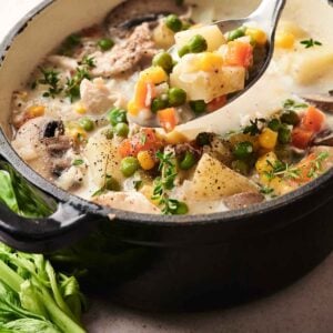 A pot of creamy chicken pot pie soup brimming with peas, carrots, potatoes, and herbs sits next to fresh celery on a table.