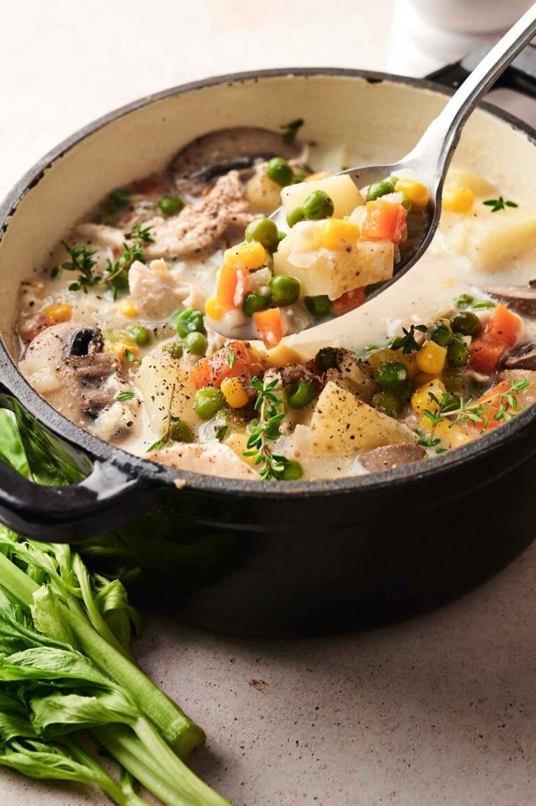 A pot of creamy chicken pot pie soup brimming with peas, carrots, potatoes, and herbs sits next to fresh celery on a table.