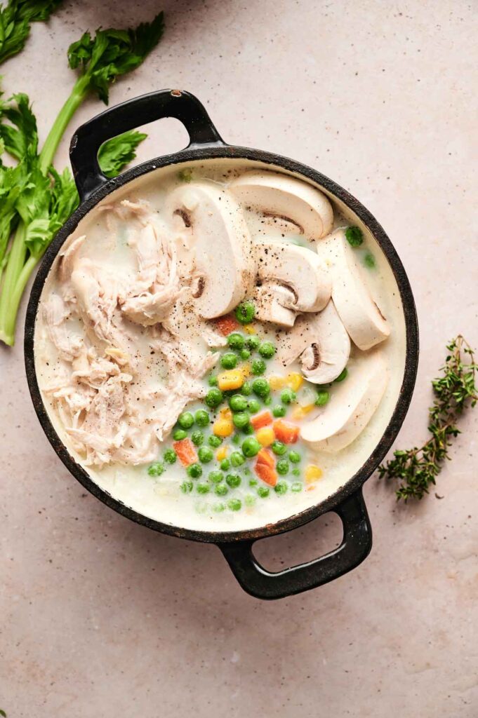 A pot with creamy chicken pot pie soup, featuring shredded chicken, sliced mushrooms, peas, and assorted vegetables. Fresh celery and thyme are nearby on a light-colored surface.