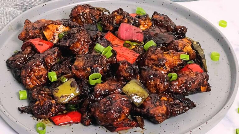 Plate of stir-fried chicken with red and green bell peppers, topped with sliced scallions, in a dark sauce.