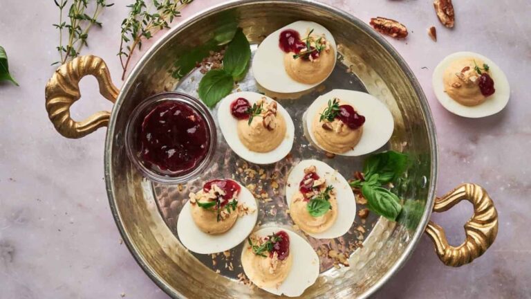 Deviled eggs topped with herbs, nuts, and cranberry sauce on a metal tray, accompanied by a small bowl of cranberry sauce and scattered herbs.