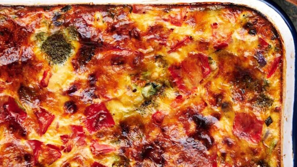 Close-up of a baked dish with a golden, crispy top, featuring melted cheese, red and green vegetables, and a browned surface.