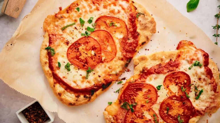 Two flatbread pizzas topped with sliced tomatoes, melted cheese, and herbs on parchment paper.