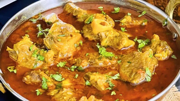 A bowl of goat curry in rich red gravy, garnished with chopped cilantro.