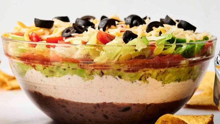 A layered dip with black beans, sour cream, guacamole, salsa, lettuce, cheese, tomatoes, and black olives in a clear bowl.