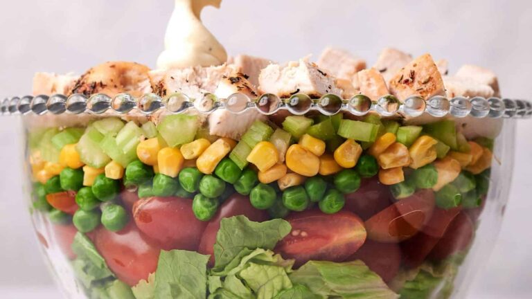 Layered salad in a glass bowl with lettuce, cherry tomatoes, peas, corn, celery, and grilled chicken, topped with a dollop of mayonnaise.