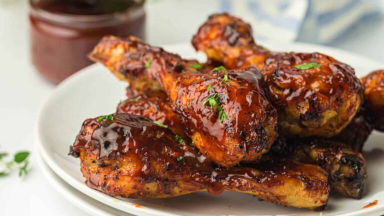 Grilled chicken drumsticks with barbecue sauce and herbs on a white plate, with a jar of sauce in the background.