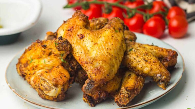 A plate of seasoned, cooked chicken wings is placed on a table near fresh cherry tomatoes.