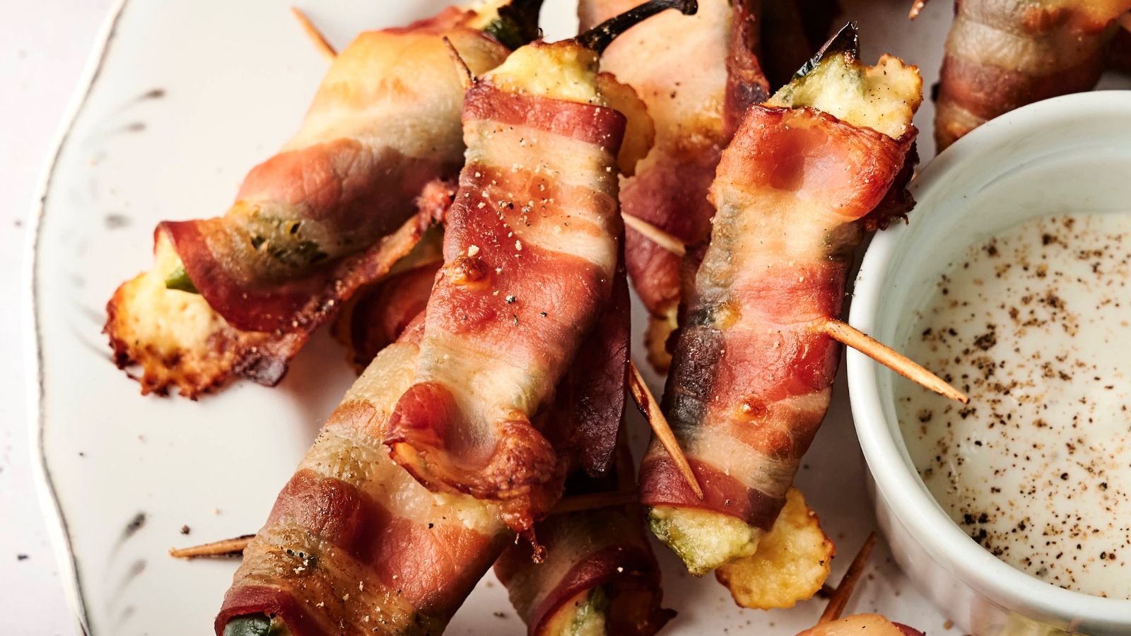 Bacon-wrapped jalapeños filled with cheese, secured with toothpicks, on a plate next to a small bowl of dipping sauce.