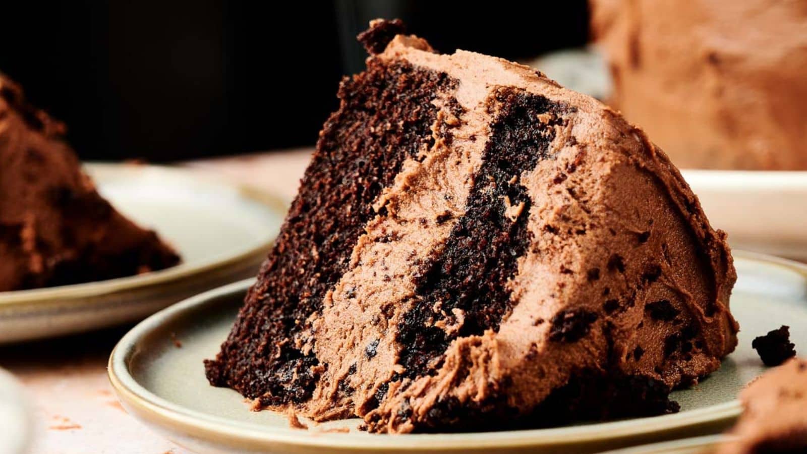 A slice of chocolate cake with rich frosting sits on a light-colored plate.