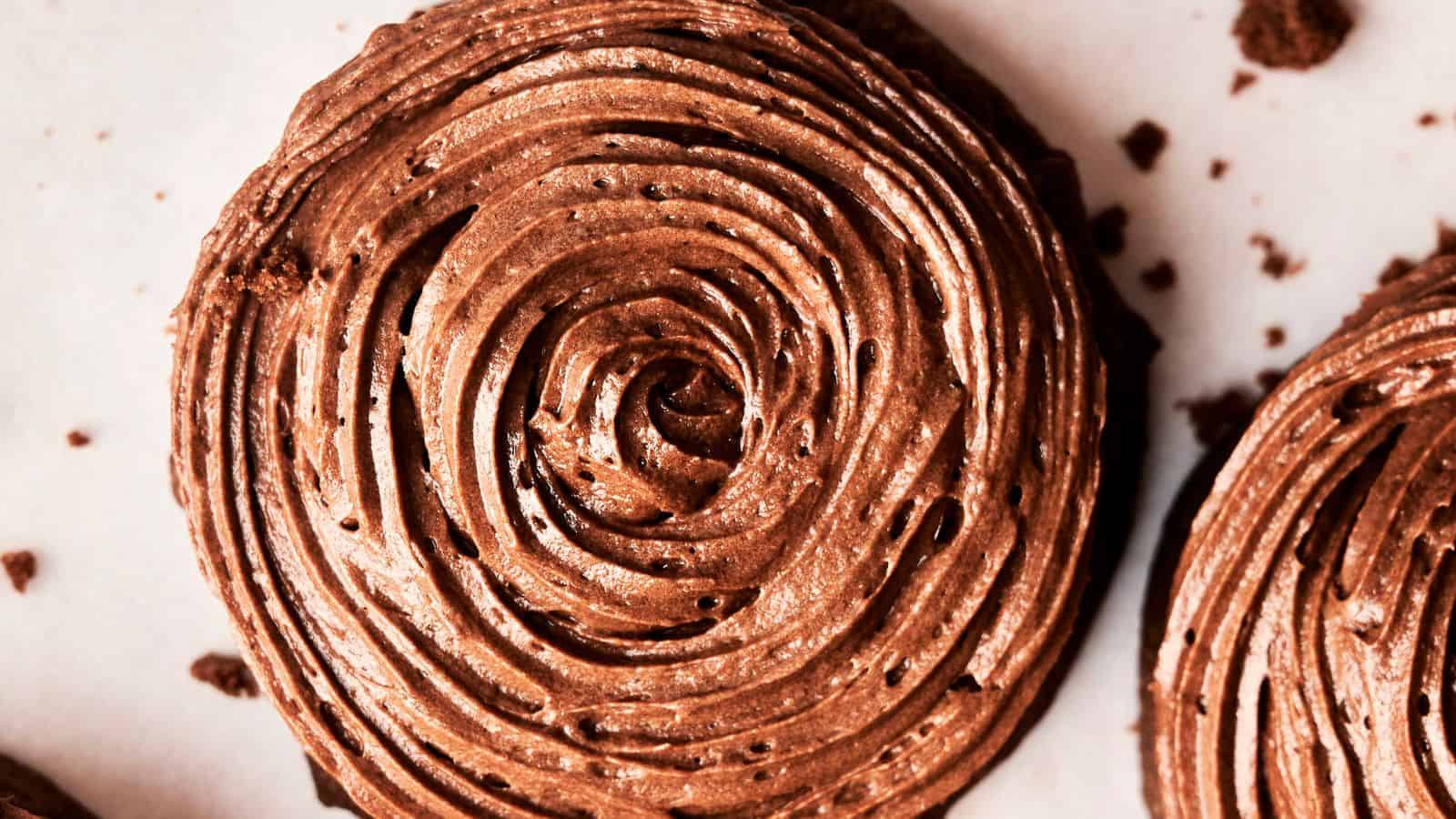 A close-up of a chocolate frosted cookie with swirl icing on a white surface, reminiscent of Copycat Crumbl Chocolate Cake Cookies and surrounded by crumbs.