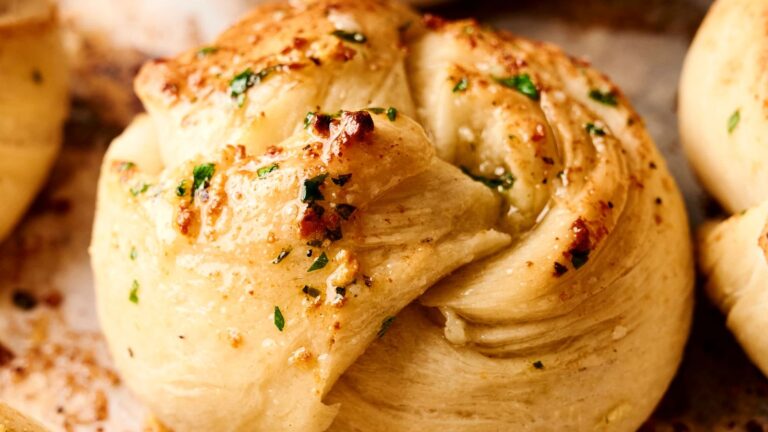 Close-up of a golden-brown garlic knot topped with herbs and seasoning on a baking tray.