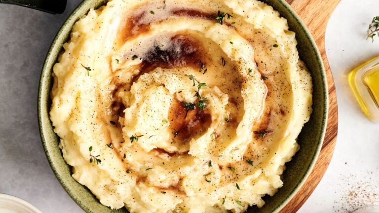 A bowl of mashed potatoes topped with gravy and garnished with herbs, placed on a wooden board.