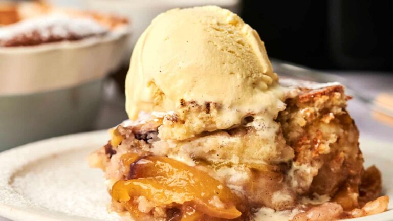 A slice of peach cobbler topped with a scoop of vanilla ice cream on a white plate.