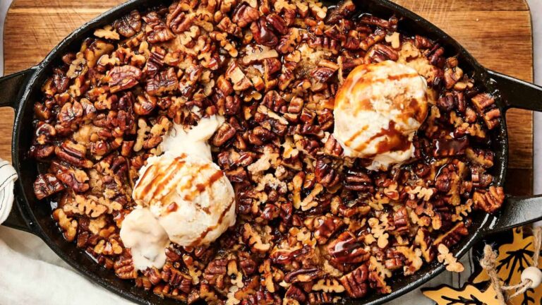 A skillet filled with a pecan dessert topped with two scoops of vanilla ice cream and caramel drizzle.