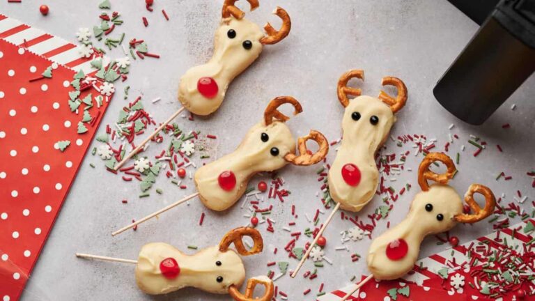 Five reindeer-themed pops made of cookies with pretzel antlers, chocolate eyes, and red noses, surrounded by festive sprinkles on a countertop.