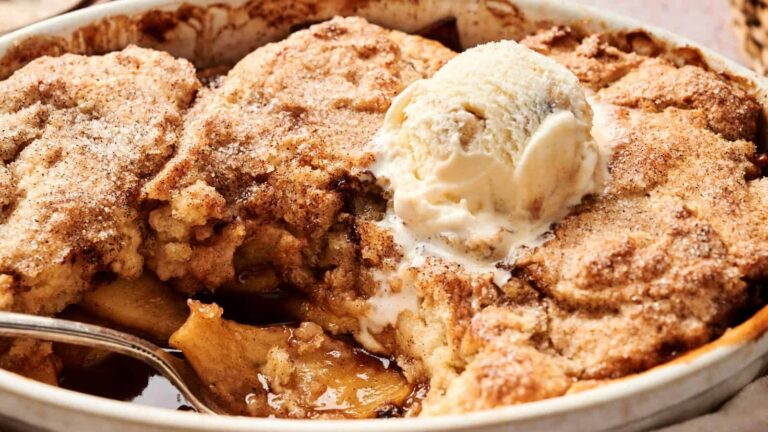 Apple cobbler topped with a scoop of vanilla ice cream in a baking dish. A serving spoon rests in the dish.