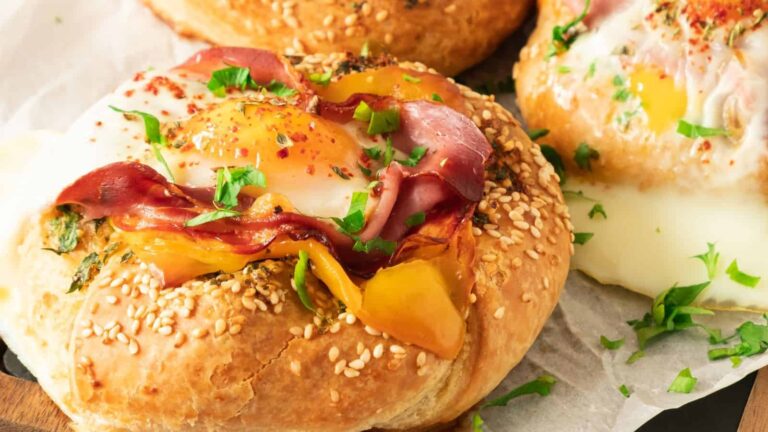 A sesame seed bun topped with a baked egg, slices of meat, bell pepper, and herbs.