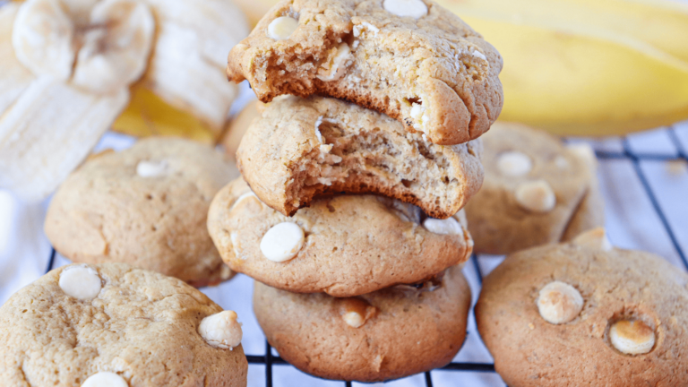 A stack of cookies with white chocolate chips, one with a bite taken out, on a cooling rack with banana slices in the background.