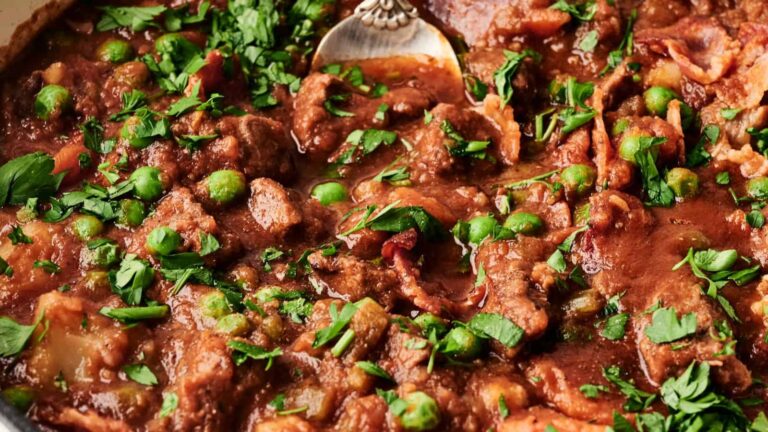 Close-up of a hearty stew with chunks of meat, peas, and vibrant green herbs in a rich sauce, with a spoon partially submerged in the mixture.