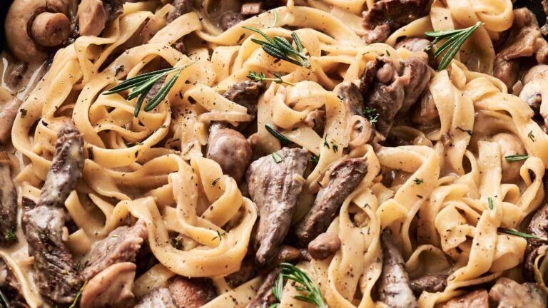Close-up of fettuccine pasta mixed with slices of beef, mushrooms, and garnished with rosemary sprigs.