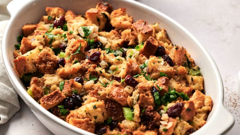 A white dish filled with baked stuffing, featuring cubes of bread, chopped herbs, celery, onion, and cranberries.