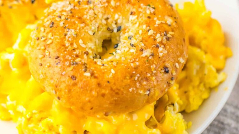 Bagel topped with seeds sits on scrambled eggs and melted cheese on a plate.