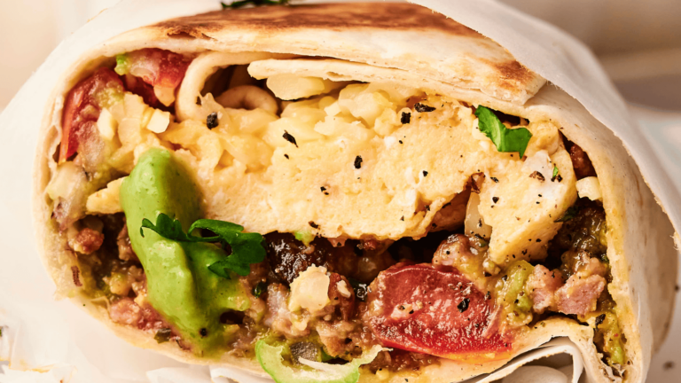 Close-up of a breakfast burrito with eggs, tomatoes, avocado, cheese, and herbs inside a tortilla.