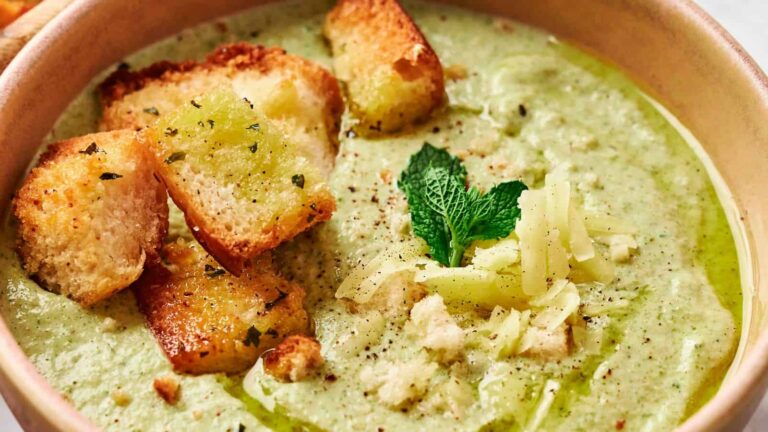 A bowl of green soup garnished with croutons, shredded cheese, a mint leaf, and a drizzle of oil.