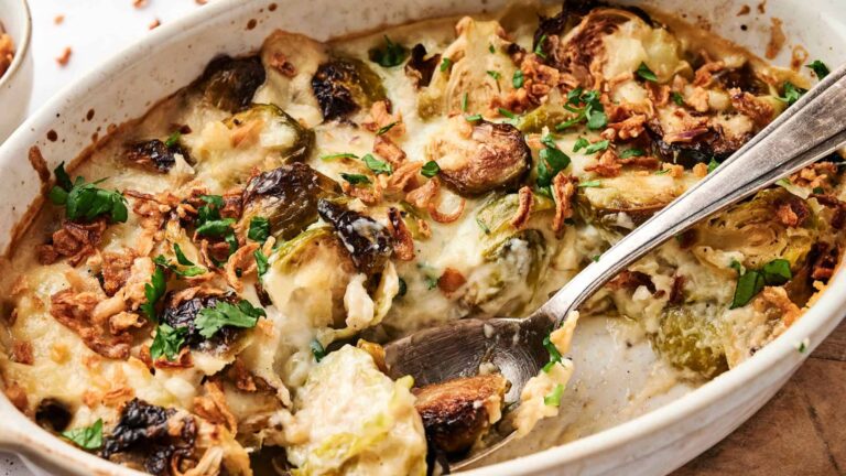 A baked casserole with Brussels sprouts, creamy sauce, and a topping of chopped herbs and crispy onions. A serving spoon rests in the dish.