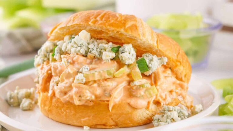 A flaky croissant filled with creamy chicken salad topped with blue cheese crumbles and chopped celery, served on a white plate.