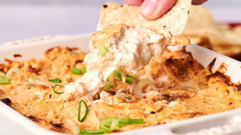 A hand dips a tortilla chip into a baked dish of creamy buffalo chicken dip, topped with sliced green onions, in a white casserole dish.