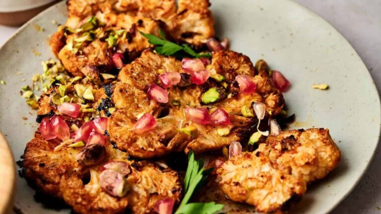 Grilled cauliflower steaks topped with pomegranate seeds and chopped pistachios on a plate, garnished with parsley.