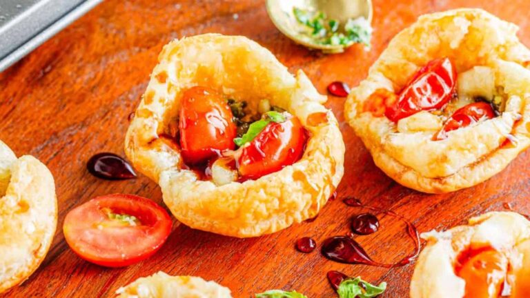 Close-up of small puff pastry cups filled with cherry tomatoes, herbs, and a dark sauce on a wooden surface.