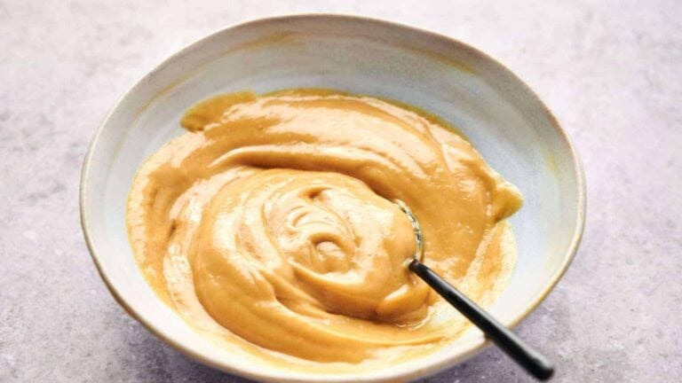 A bowl of smooth, creamy peanut butter with a spoon inside, on a light gray surface.