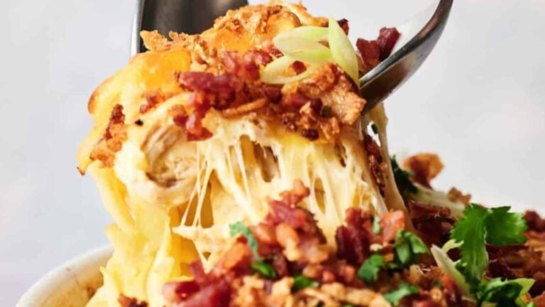 A spoon lifts a cheesy, baked pasta dish topped with crumbled bacon and garnished with green onions and cilantro.