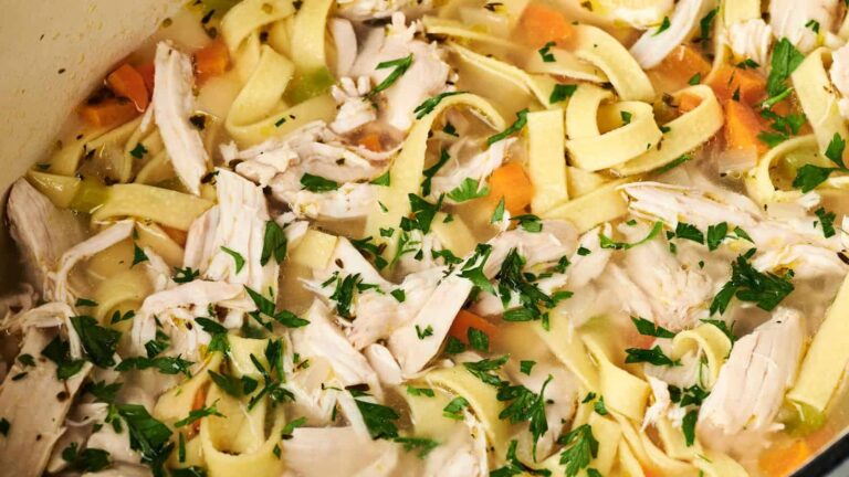 A close-up of chicken noodle soup with shredded chicken, egg noodles, carrots, and parsley in a broth.