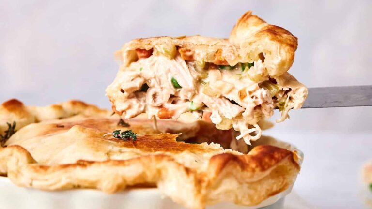 A slice of chicken pot pie with a flaky crust on a spatula, showing the creamy filling with chicken, vegetables, and herbs.
