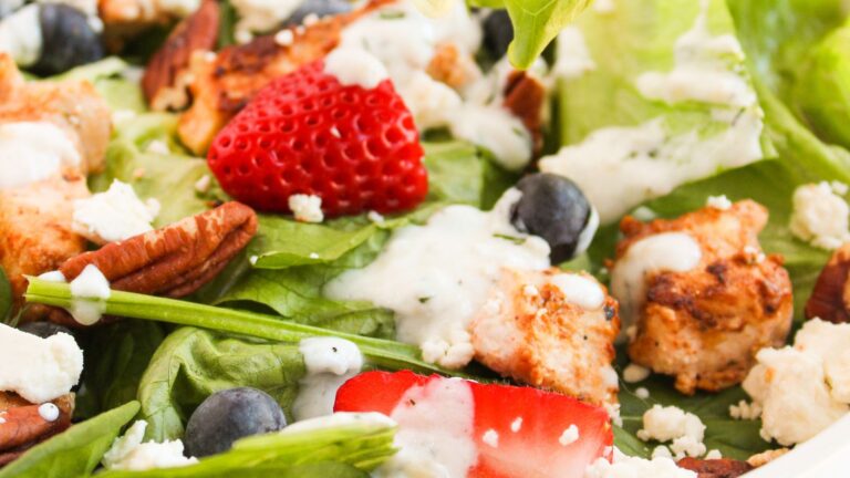 Close-up of a salad with grilled chicken, strawberries, blueberries, pecans, spinach, and crumbled cheese, topped with creamy dressing.