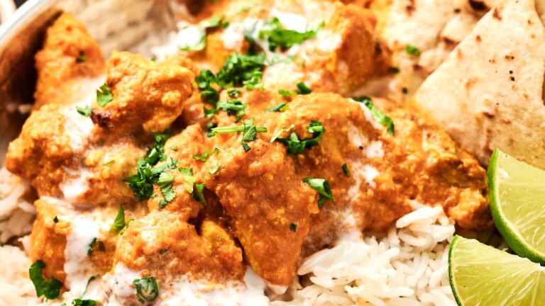 A dish of chicken tikka masala on rice, garnished with chopped herbs and served with lime wedges and naan bread.