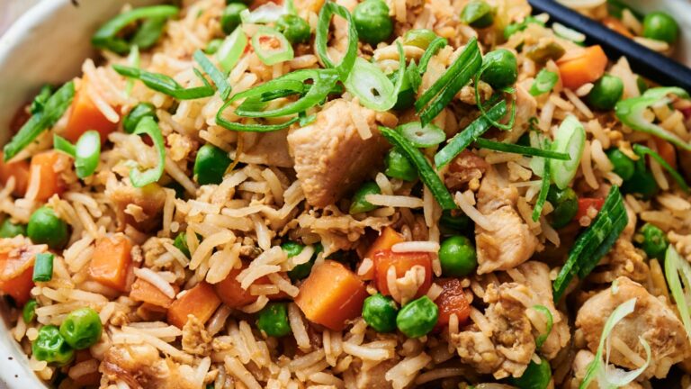 Close-up of a bowl of fried rice with chicken, peas, carrots, and sliced green onions. Black chopsticks rest on the side.